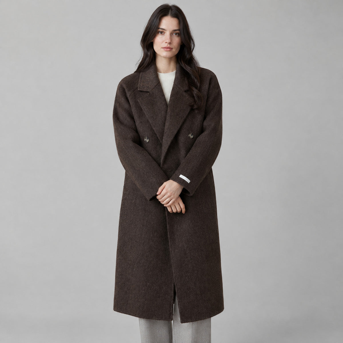 Dark brown double-breasted long wool coat worn by a model against a neutral studio backdrop.